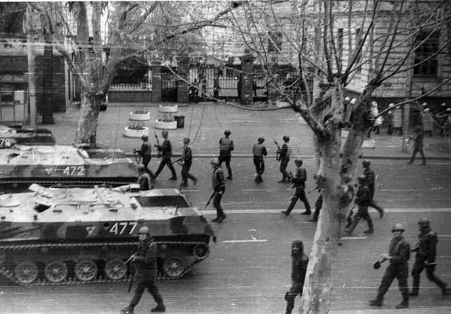 A black and white photograph of soldiers walking beside armoured vehicles on a tree-lined city street